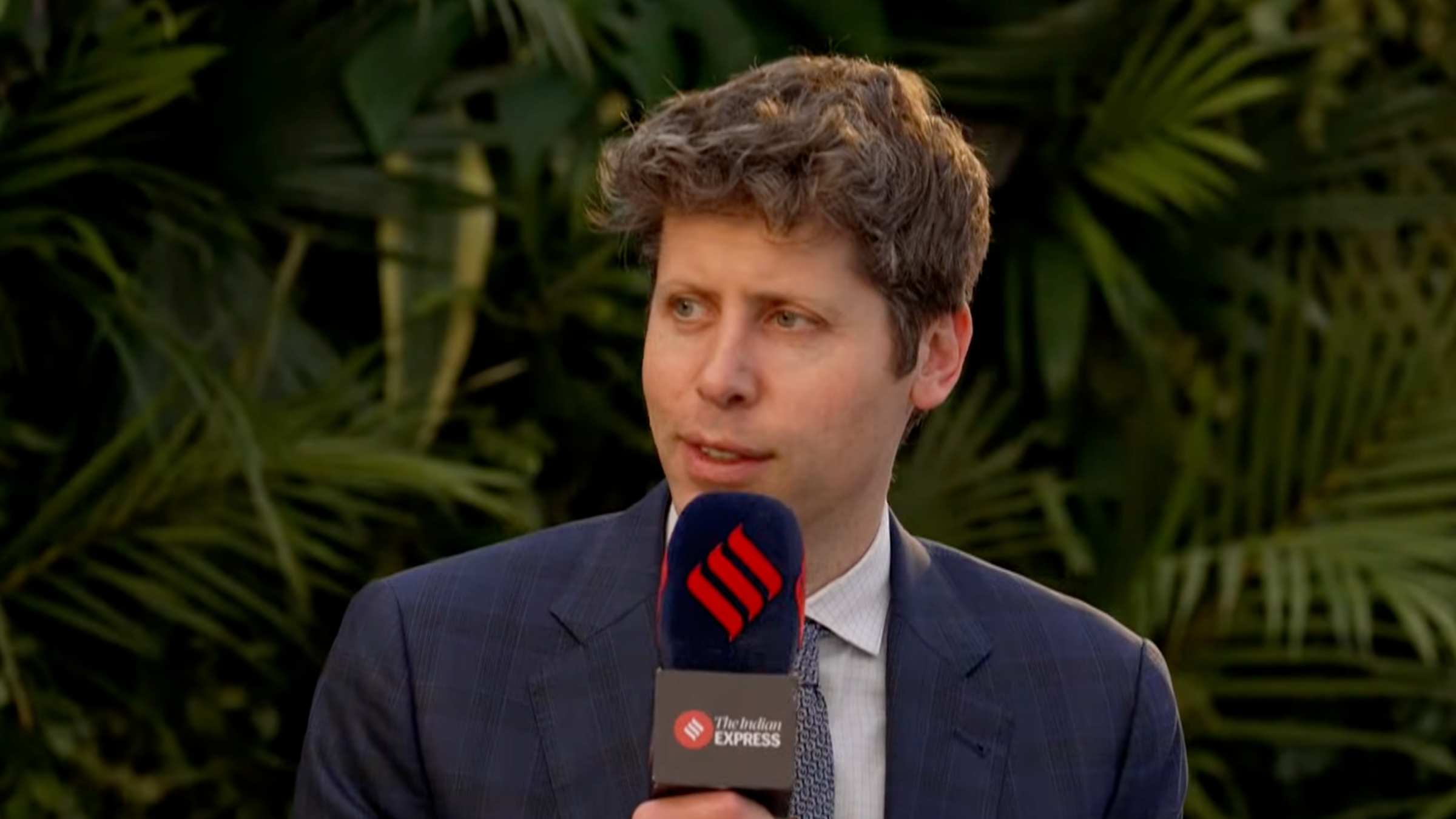 Sam Altman holding a microphone with ferns in the background